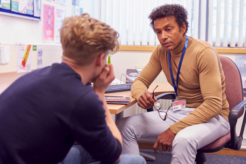 Male counselor sits on an office chair speaking to a young person